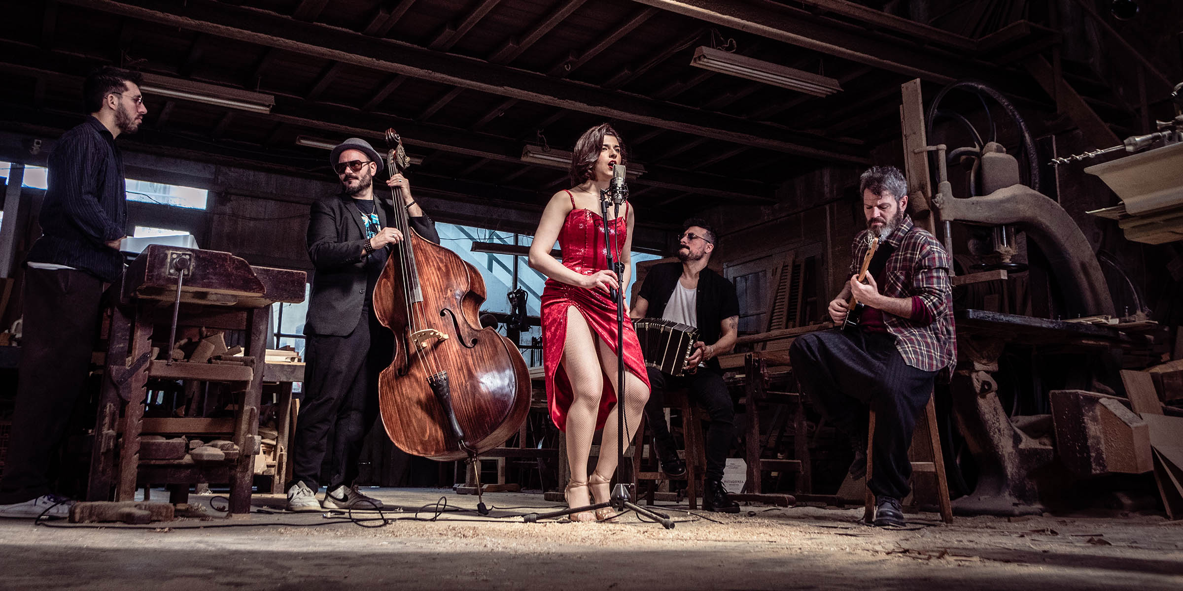 Fem personer i et band i et sløydverksted. Vokalist foran mikrofonen i knallrød kjole.