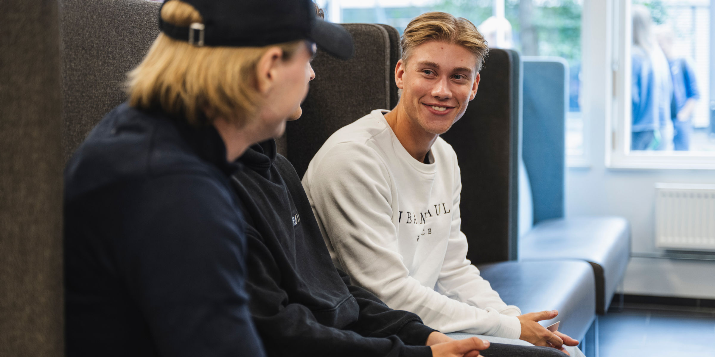 Three OsloMet students are sitting on a sofa. One of the students is smiling at the other two.