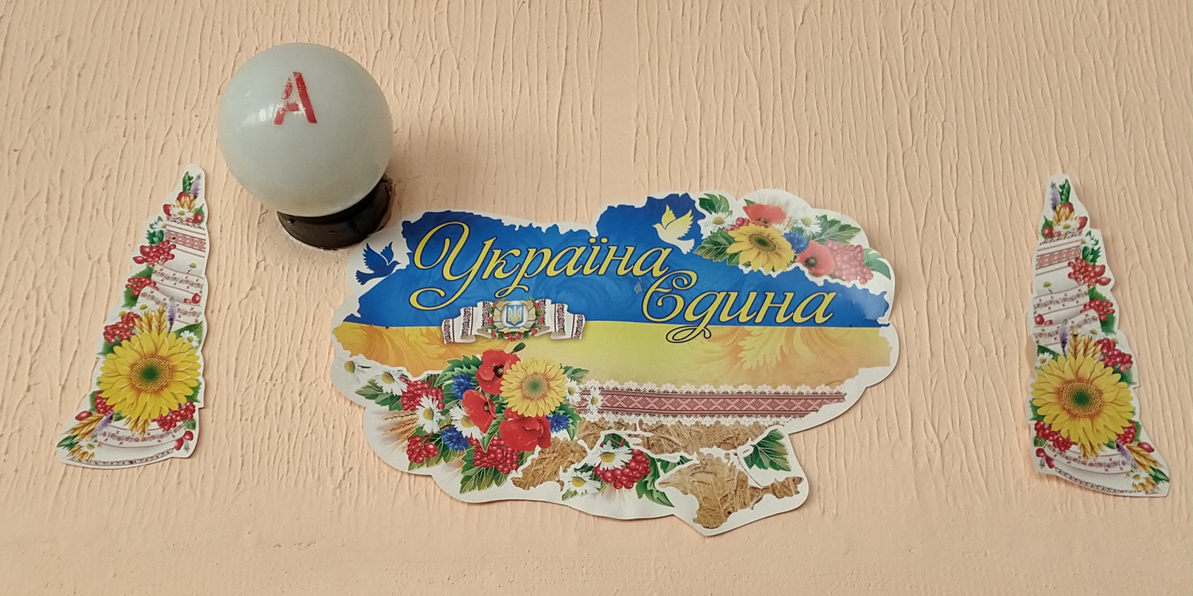 På en ferskenfarget murvegg henger en hvit lyskuppel med bokstaven A i rødt. Under kuppelen er det festet tre papirbilder, hvor en silhuett av det ukrainske kartet er midten, omgitt av dekorelementer bestående av solsikker og andre blomster.