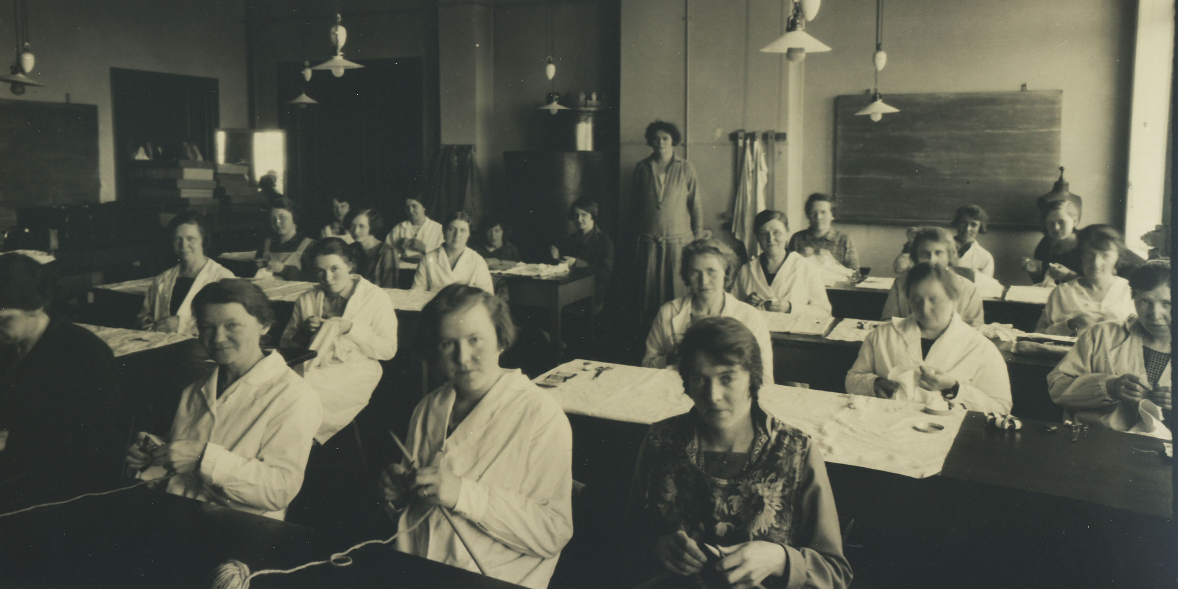 Elever ved Den kvinnelige industriskole sitter ved pultene sine og strikker.