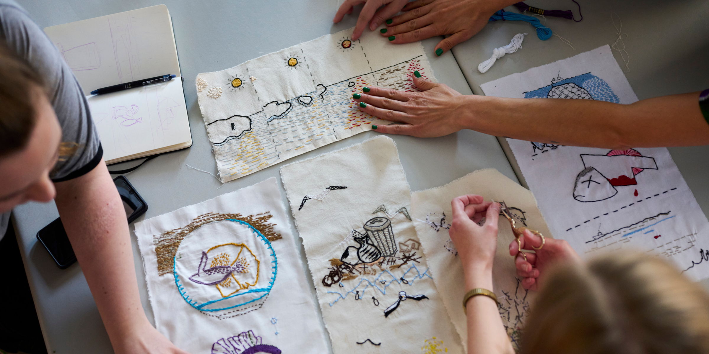 Broderier ligger på et bord og hender holder på broderiene.