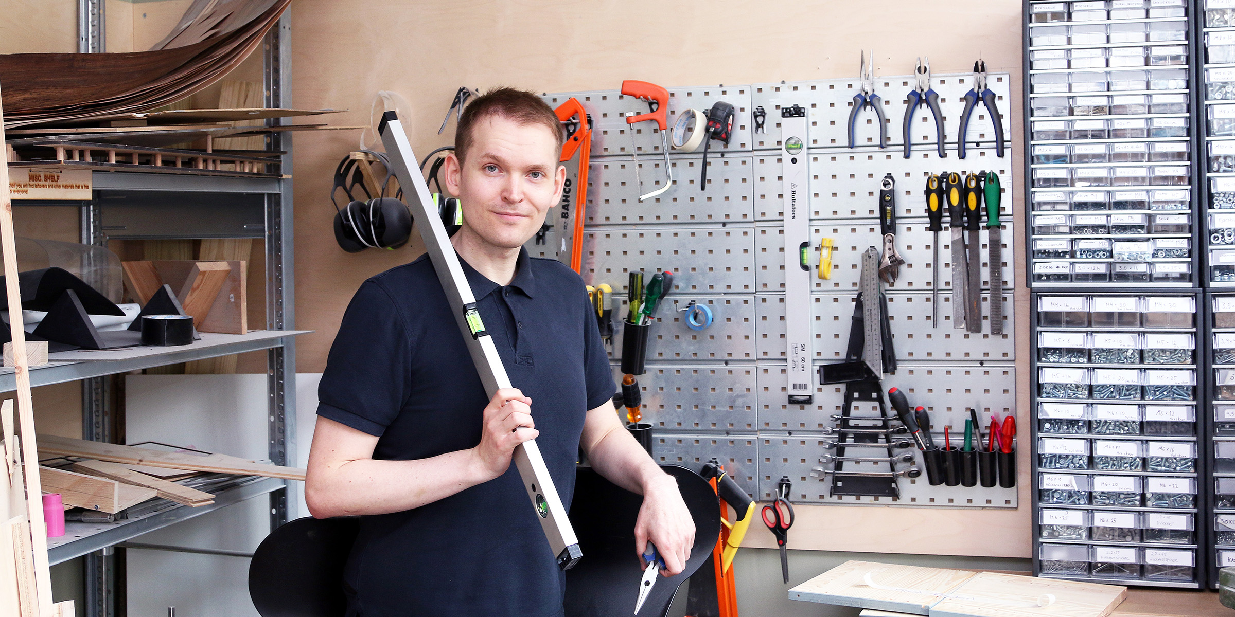 Lars with tools at OsloMet Makerspace. He has spirit levels and pliers in his hands, and on the wall behind are various tools and screws.