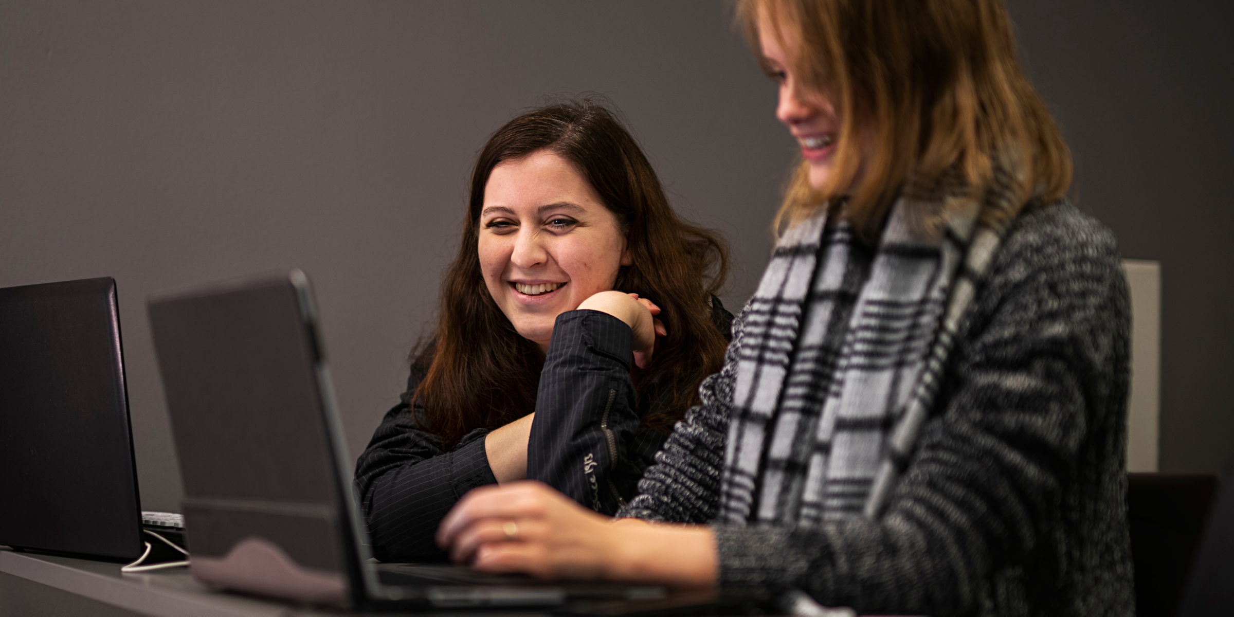 To OsloMet-studenter sitter med hver sin laptop. Begge studentene ser på den ene skjermen mens de smiler.