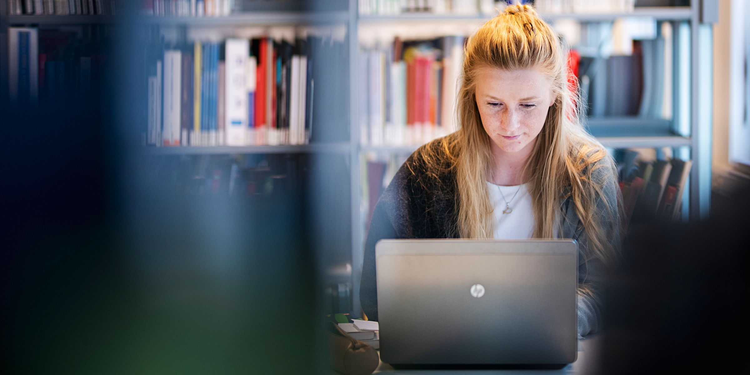 Oslomet-student sitter og ser på laptopen sin. Bak henne er en bokhylle full av bøker.