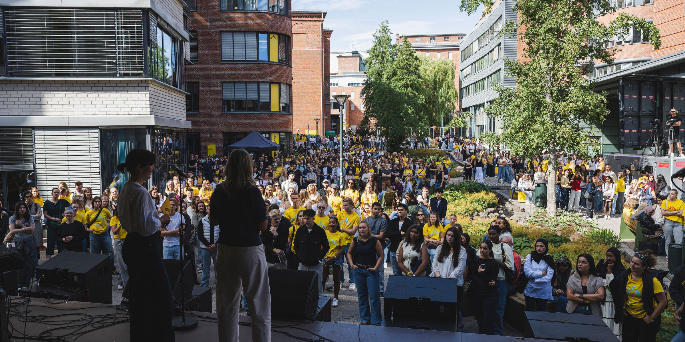 To kvinner med ryggen til kamera står på scenen med en stor folkemasse foran scenen under studiestart i 2025.