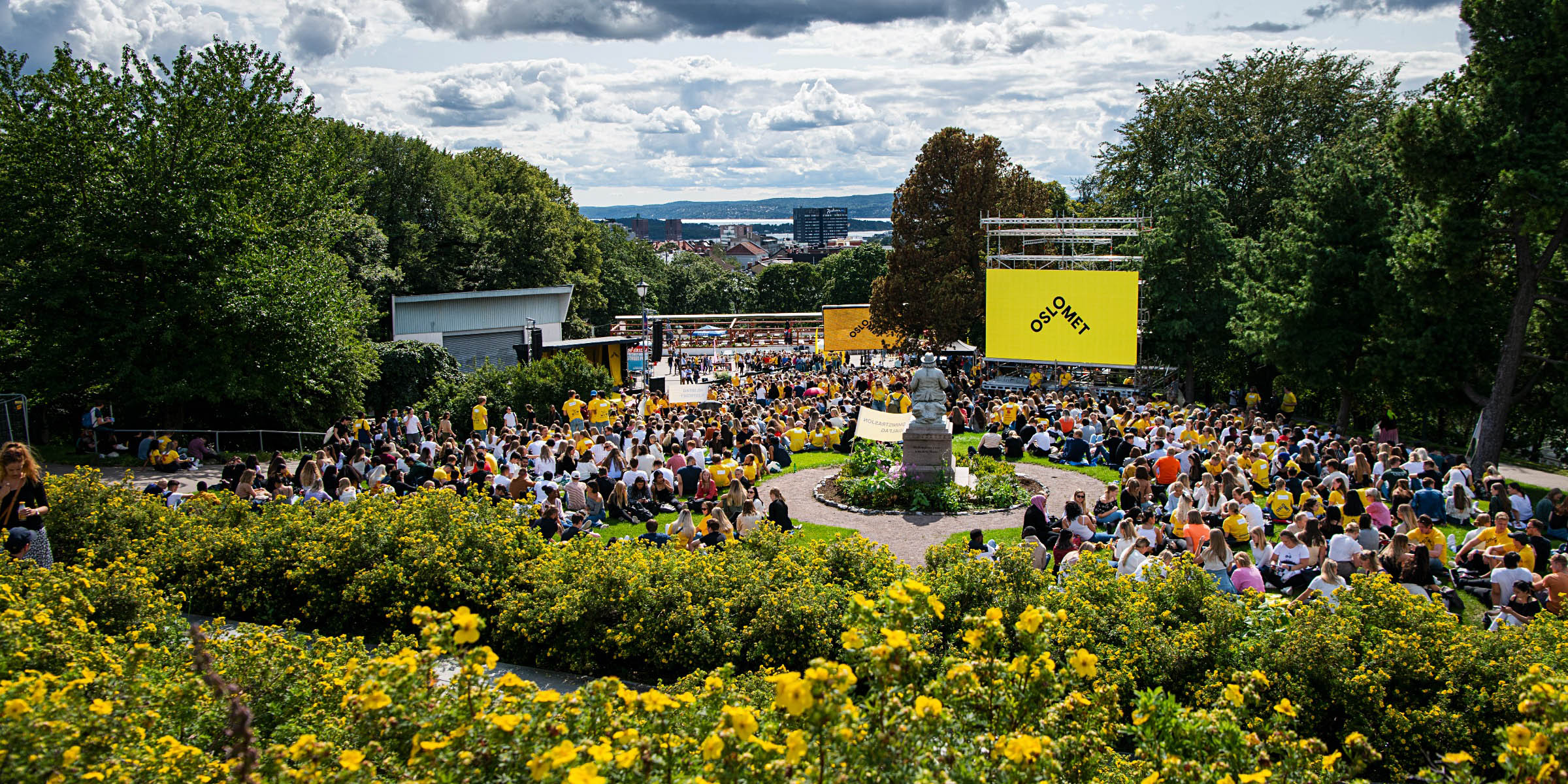 Mange mennesker i en park, gule blomster i forgrunnen. Storskjerm med OsloMet-logo på høyre side.
