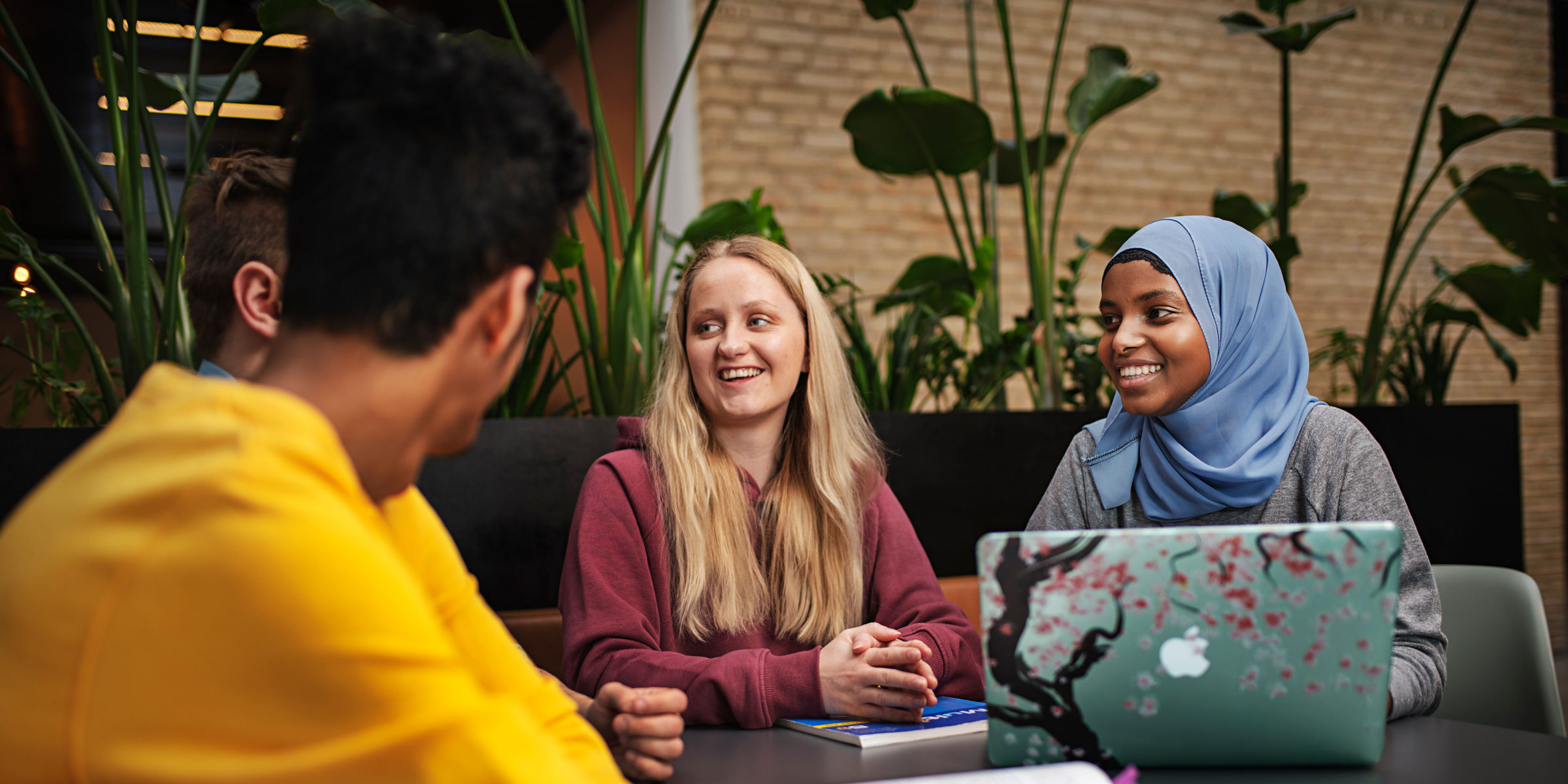 En jente med hijab og en blond jente sitter ved et bord og snakker med to mannlige studenter med ryggen til kamera.
