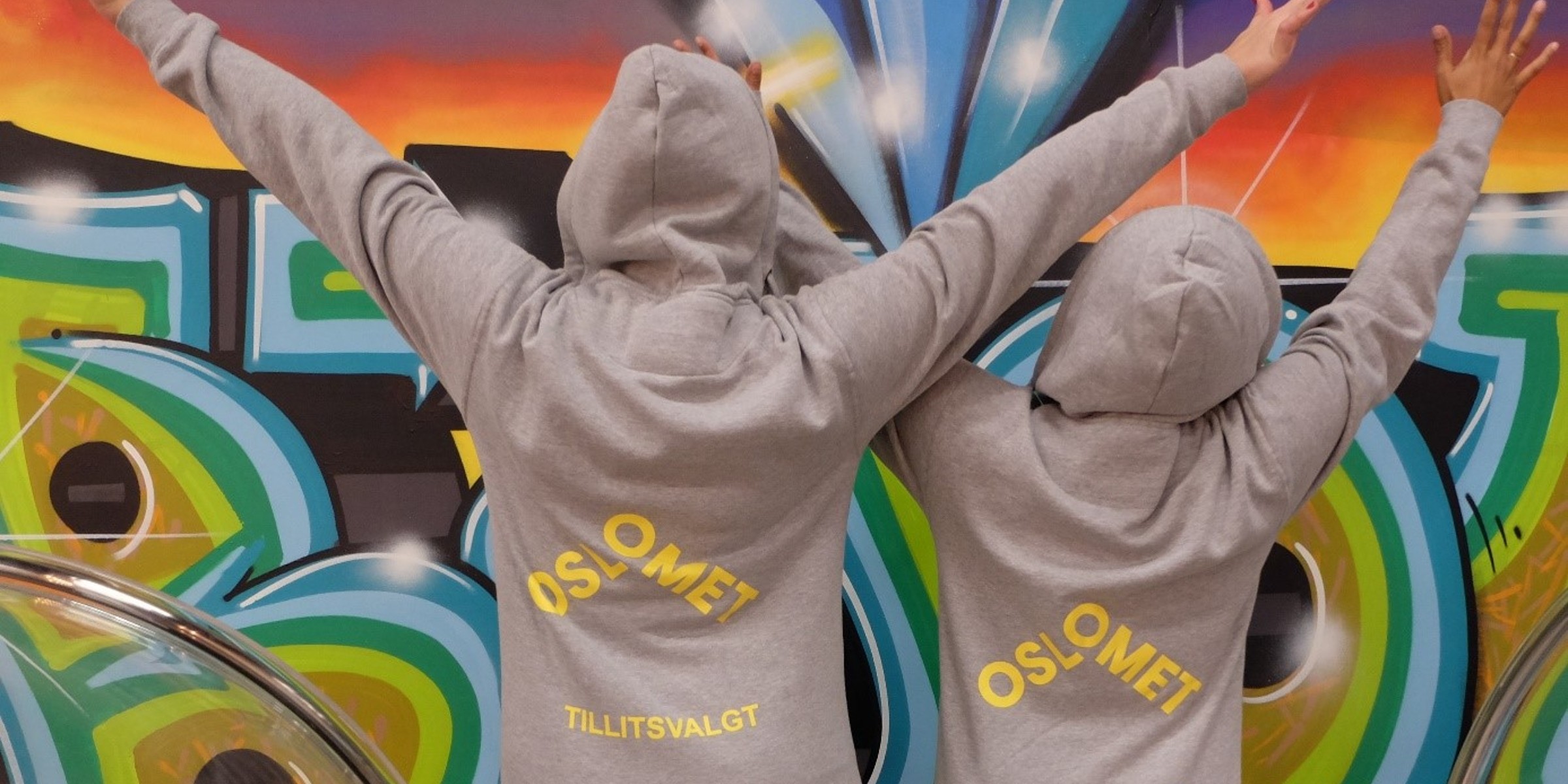 Two students stand with their backs turned in hoodies with the inscription OsloMet and Student Representative. Behind them is a graffiti wall.