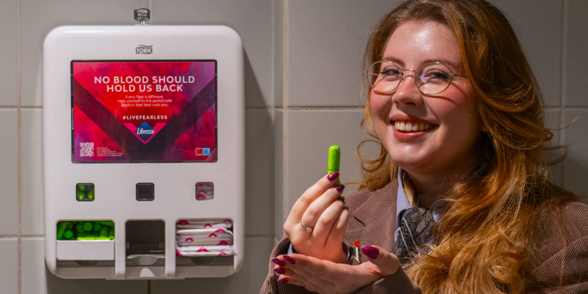 Student Parliament Leader, Victoria Skogsletten Dalen, smiles at the camera while standing next to a dispenser with pads and tampons and holding a tampon in one hand.