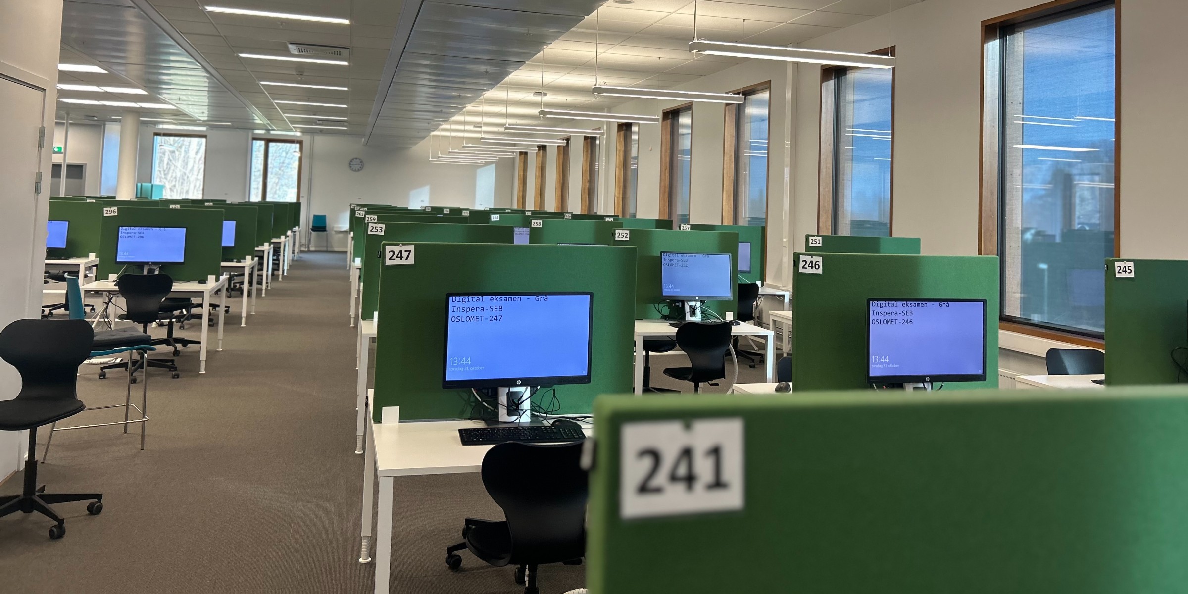 Many desks, each with its own PC, stand in a row in a large room.