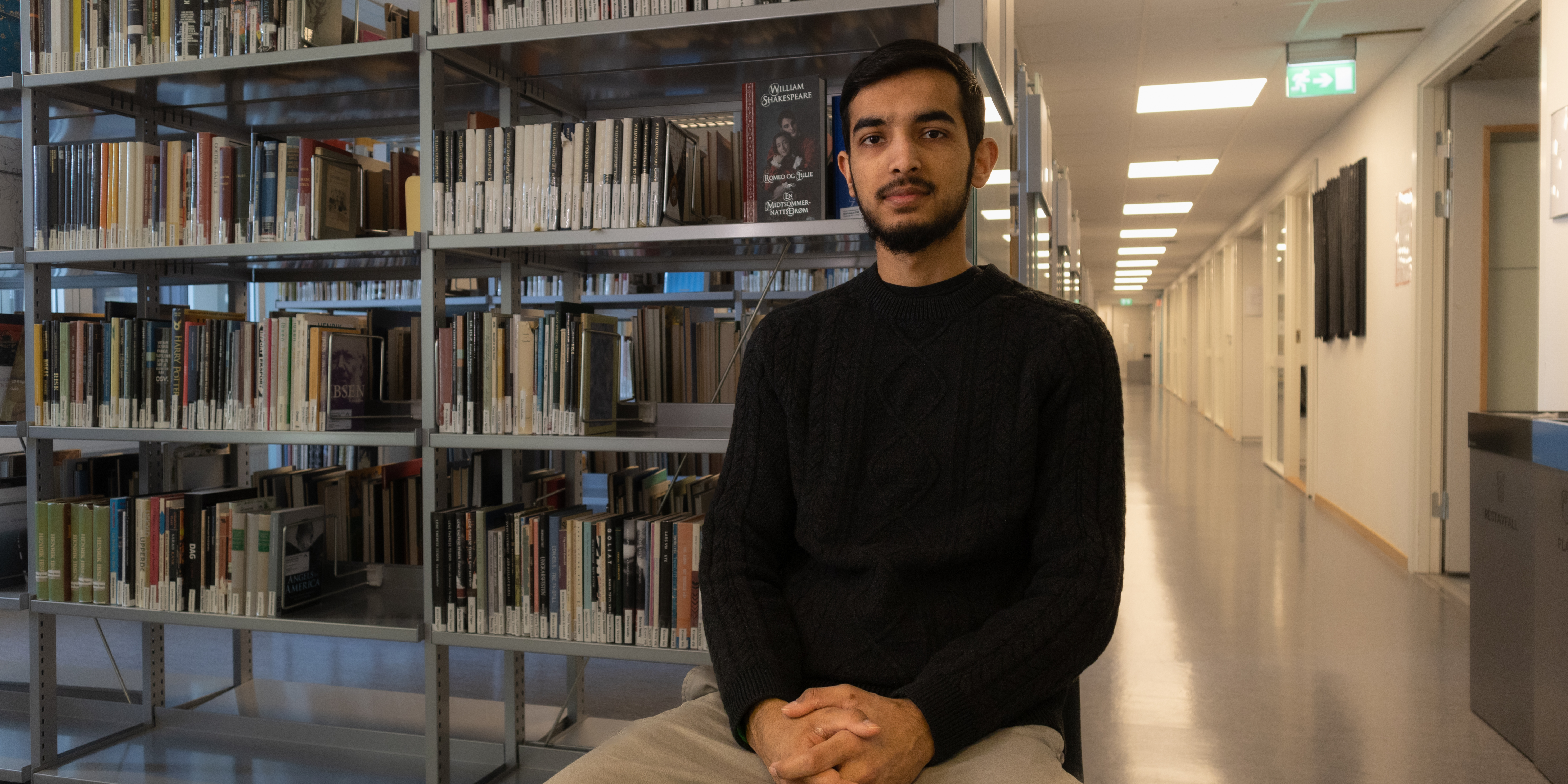 OsloMet student Mohammad Arwais Hameed sits in the university library, in front of a bookshelf.