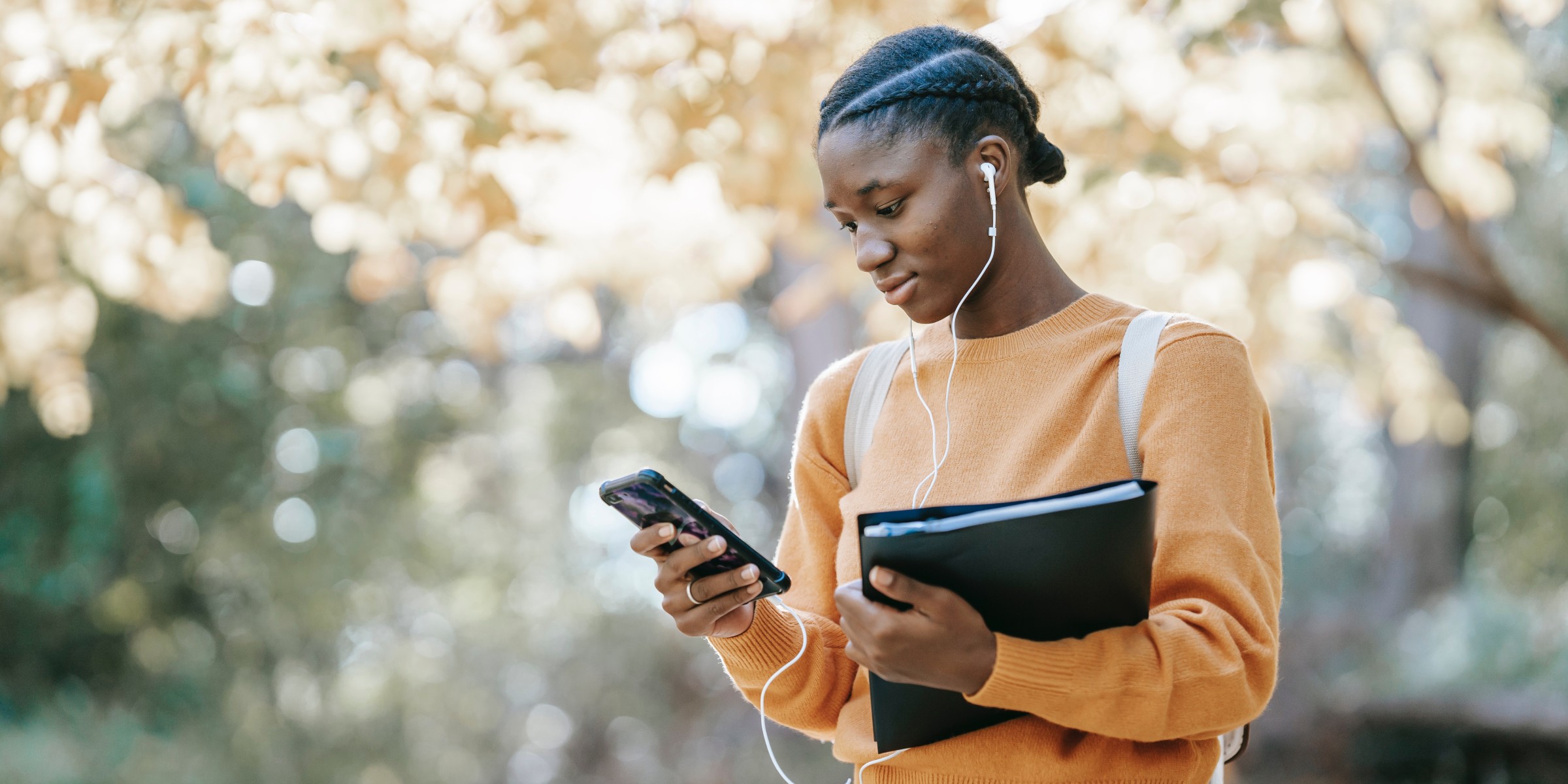 Girl with backpack holds a binder in one hand while reading on her mobile phone from the other hand.