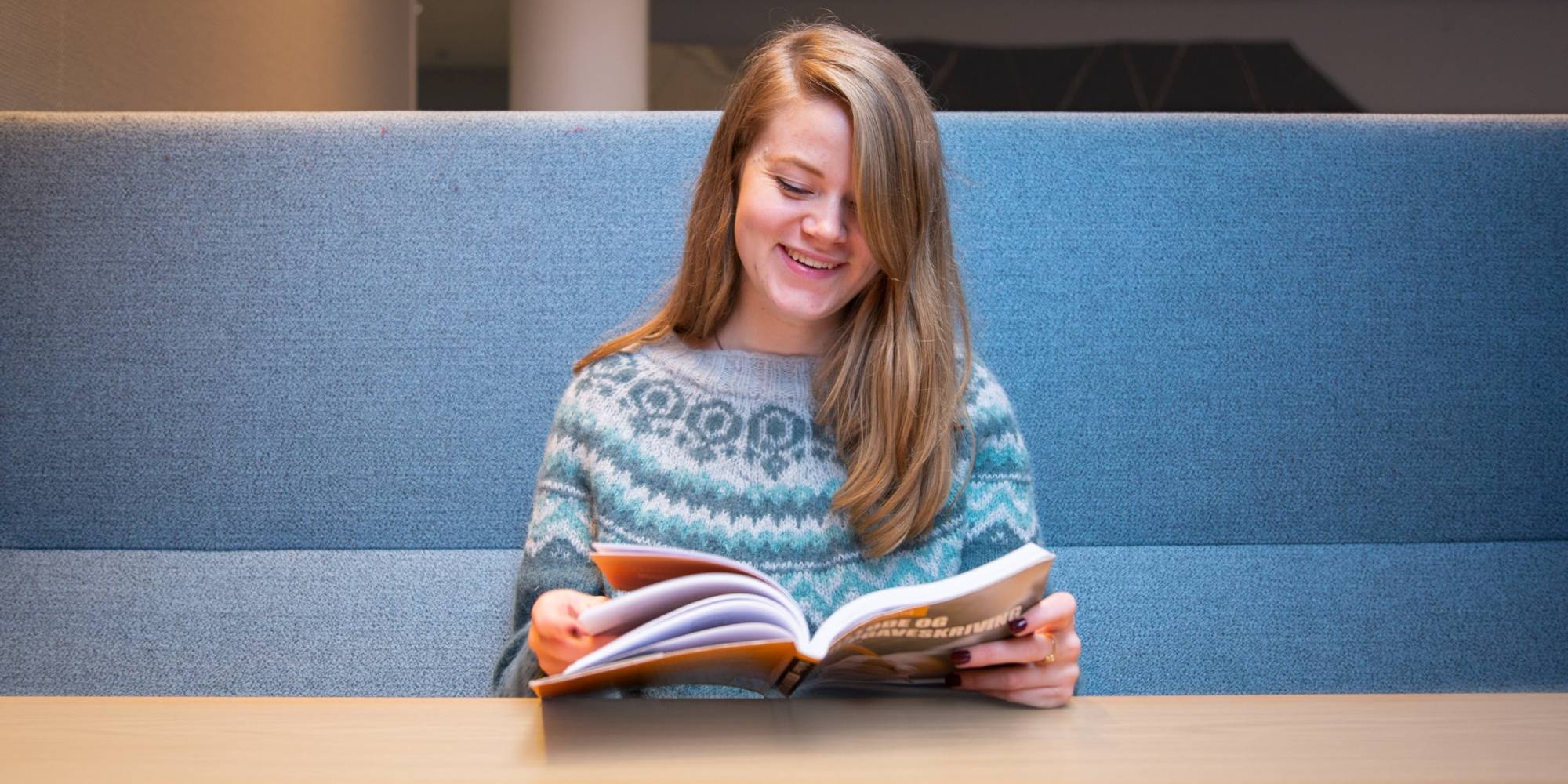 OsloMet student sitting and reading a syllabus book while smiling.