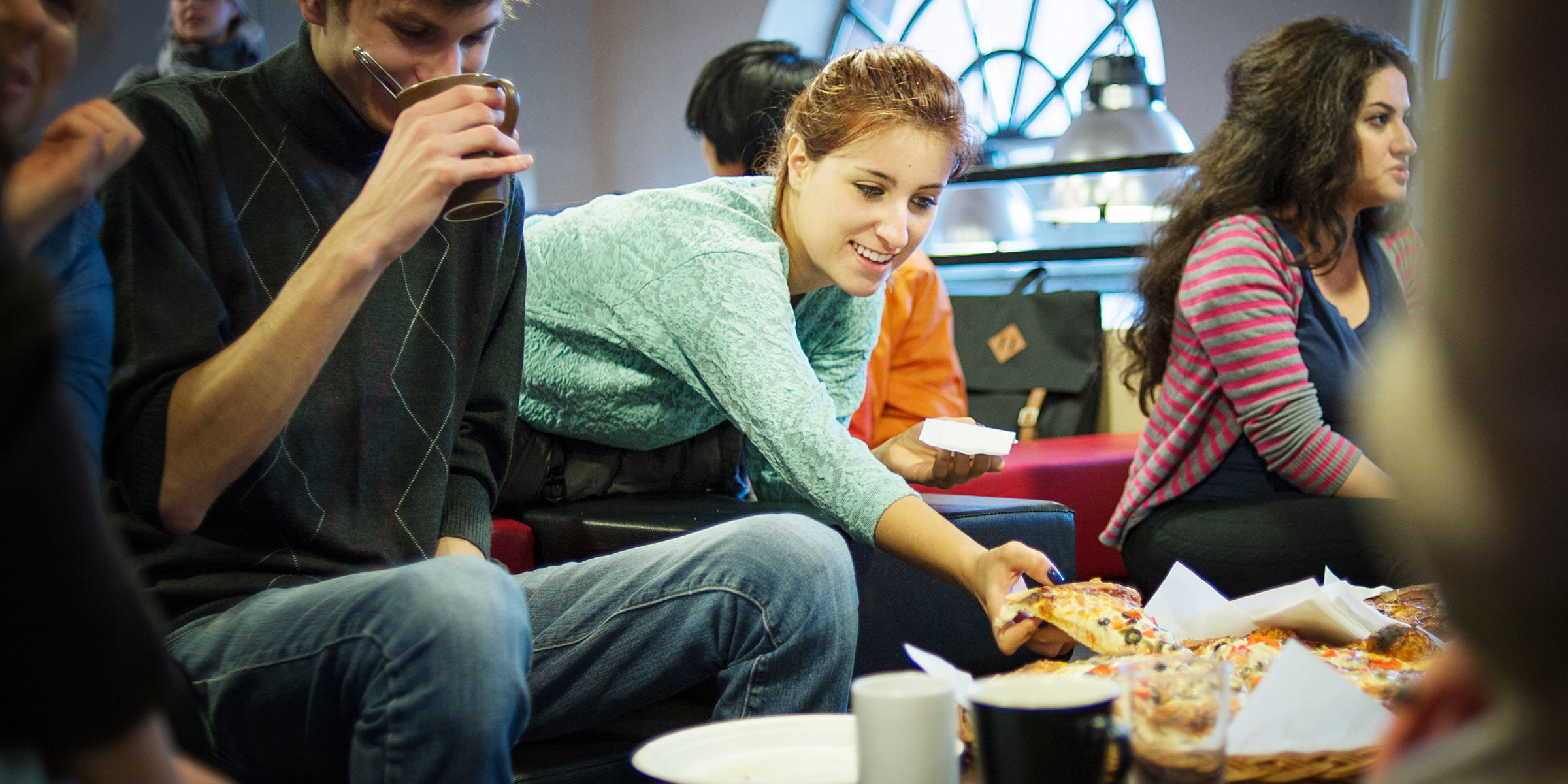 OsloMet student sits on the sofa and bends over the table to take a slice of pizza