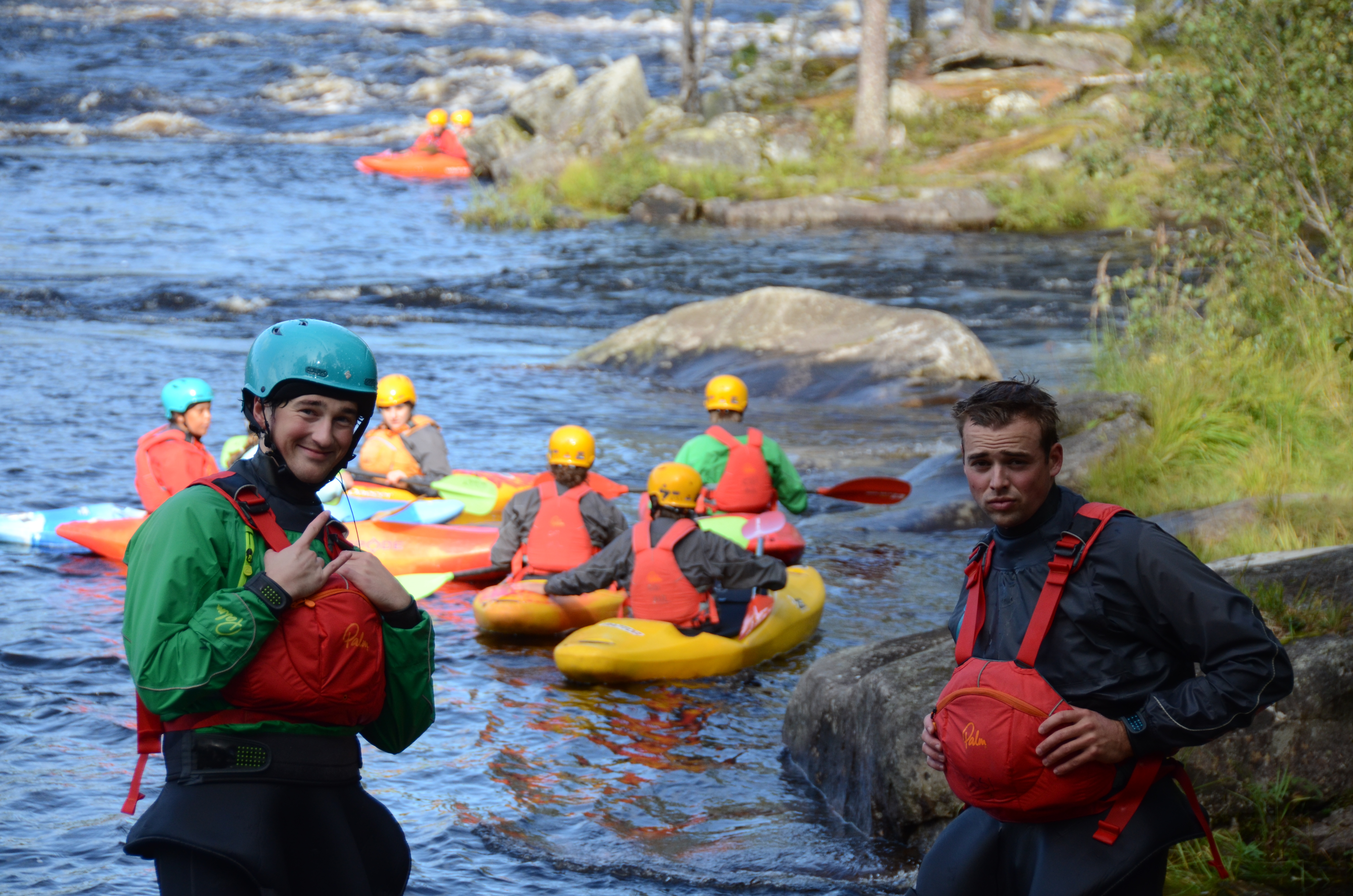 Kajakker med folk i bakgrunnen, to gutter ser inn i kamera og smiler i padleutstyr.