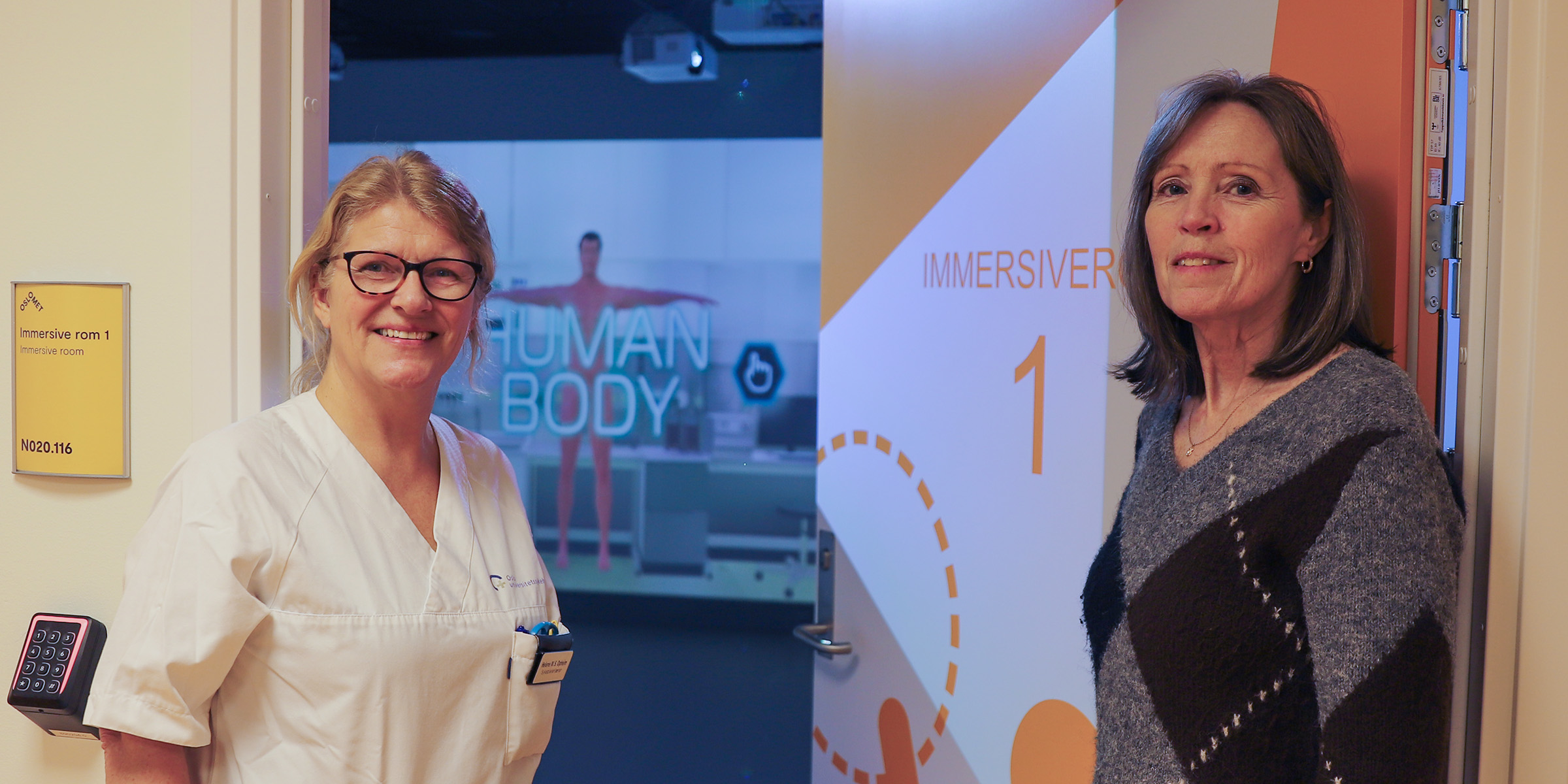 To personer står utenfor et undervisningsrom der du kan skimte en digital vegg i bakgrunnen.