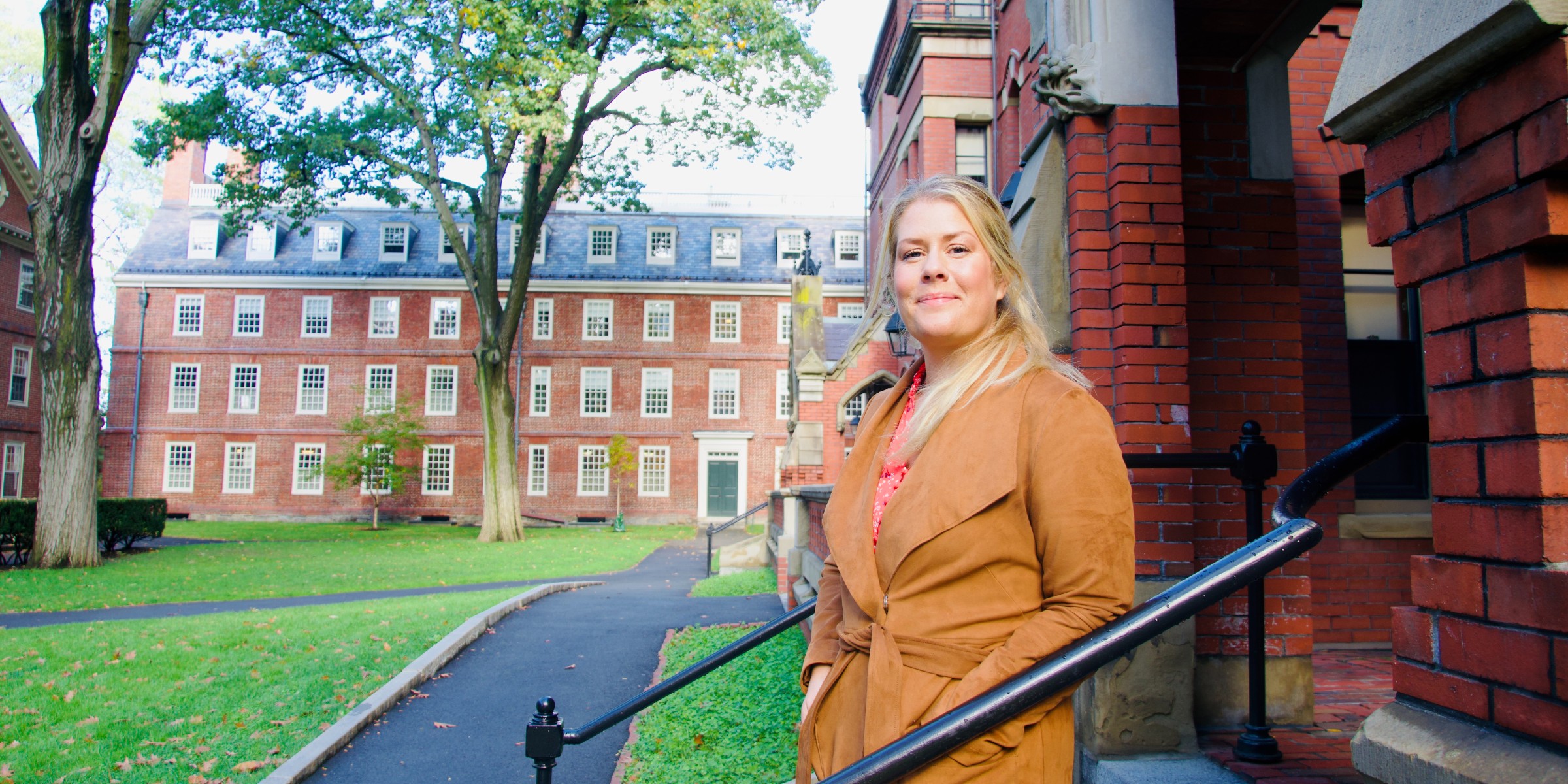 Photo of Miriam Sinkerud Johnson standing in the stairs outside a building at campus where she works. på Harvard University. Trær og bygg i bakgrunnen.