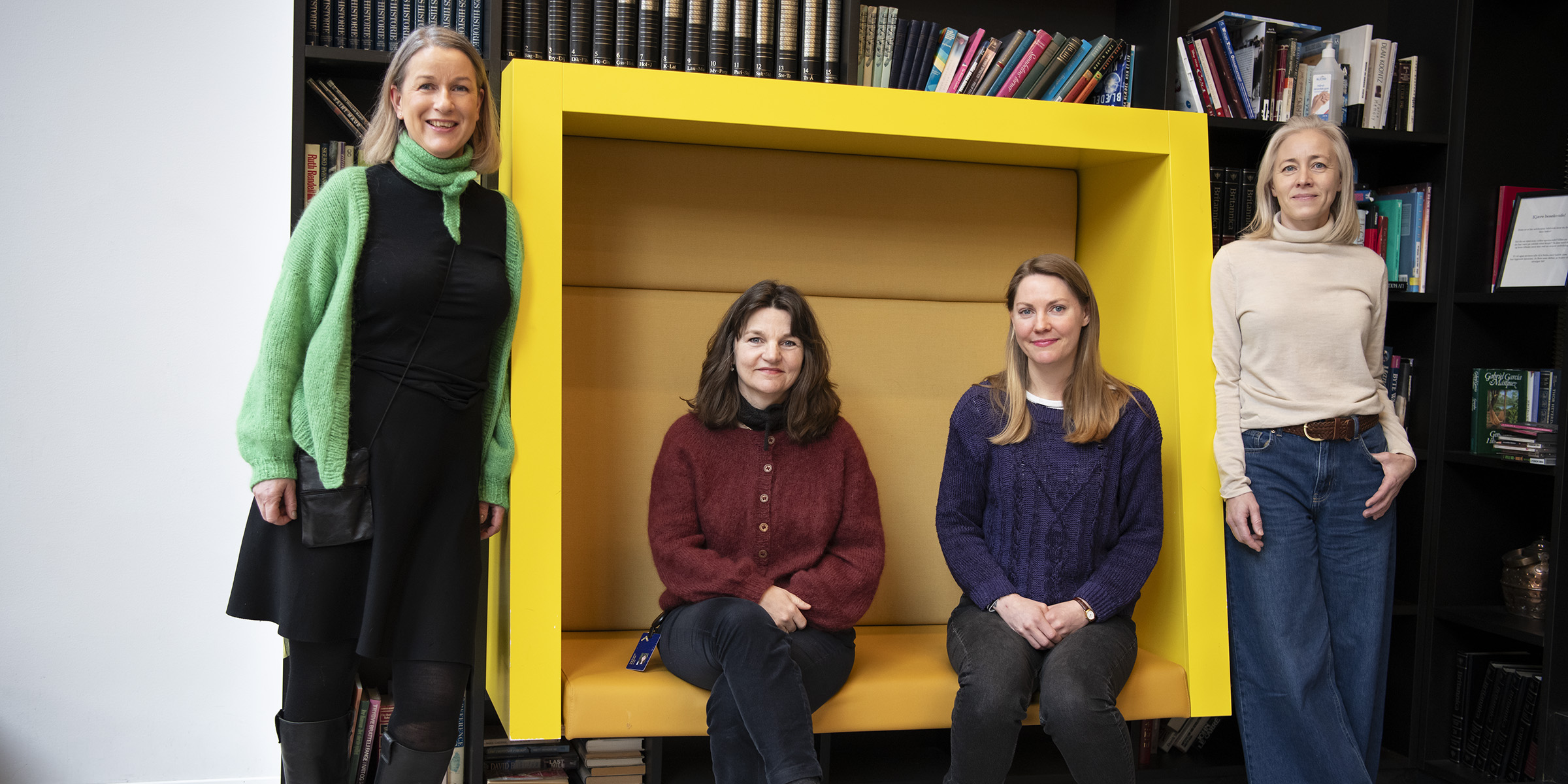 To kvinner sitter i en gul sofa, og to kvinner står på hver side av sofaen.