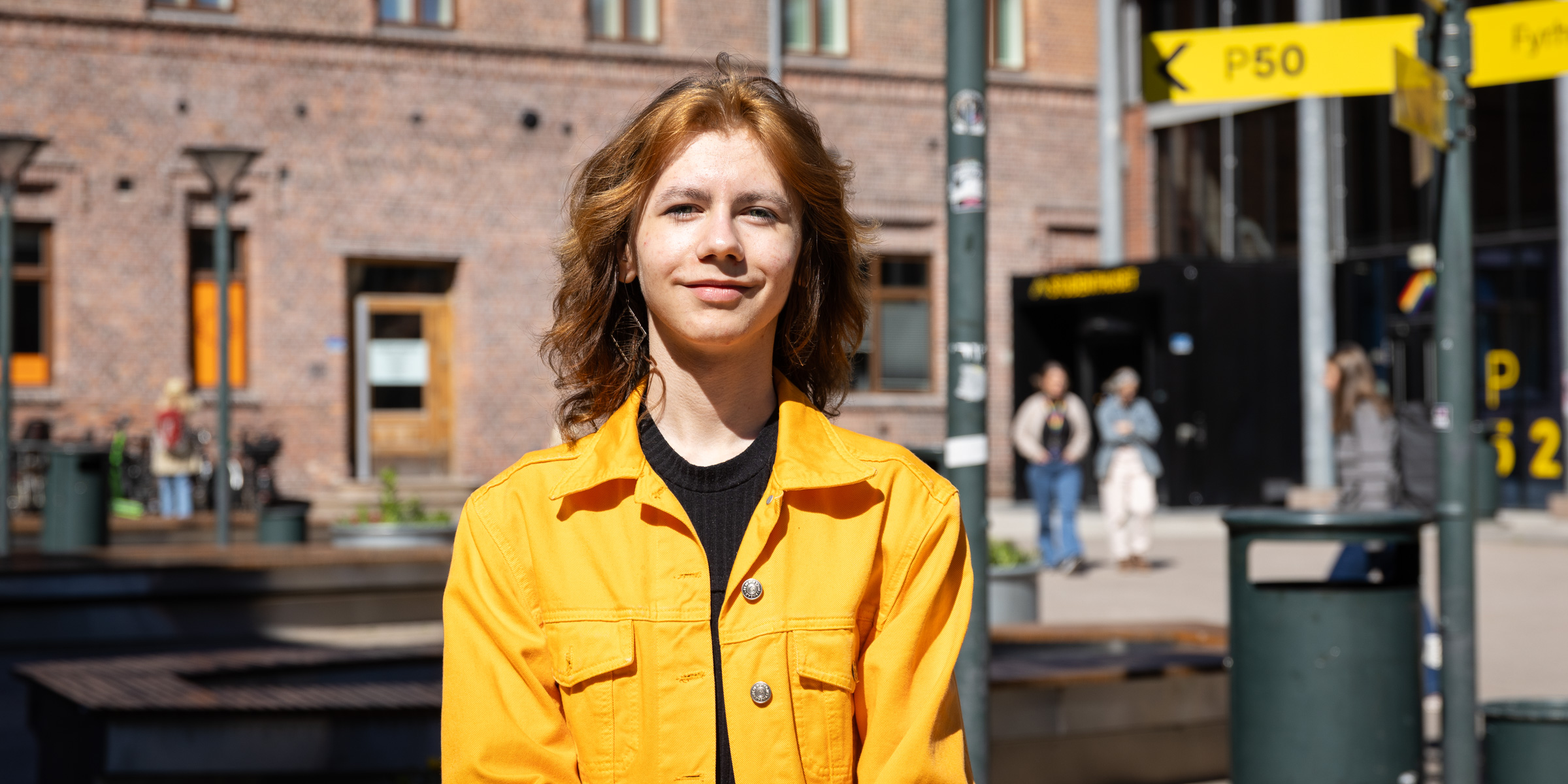 OsloMet student Dawid Sasha stands in a yellow jacket on the campus in Pilestredet and smiles at the camera.