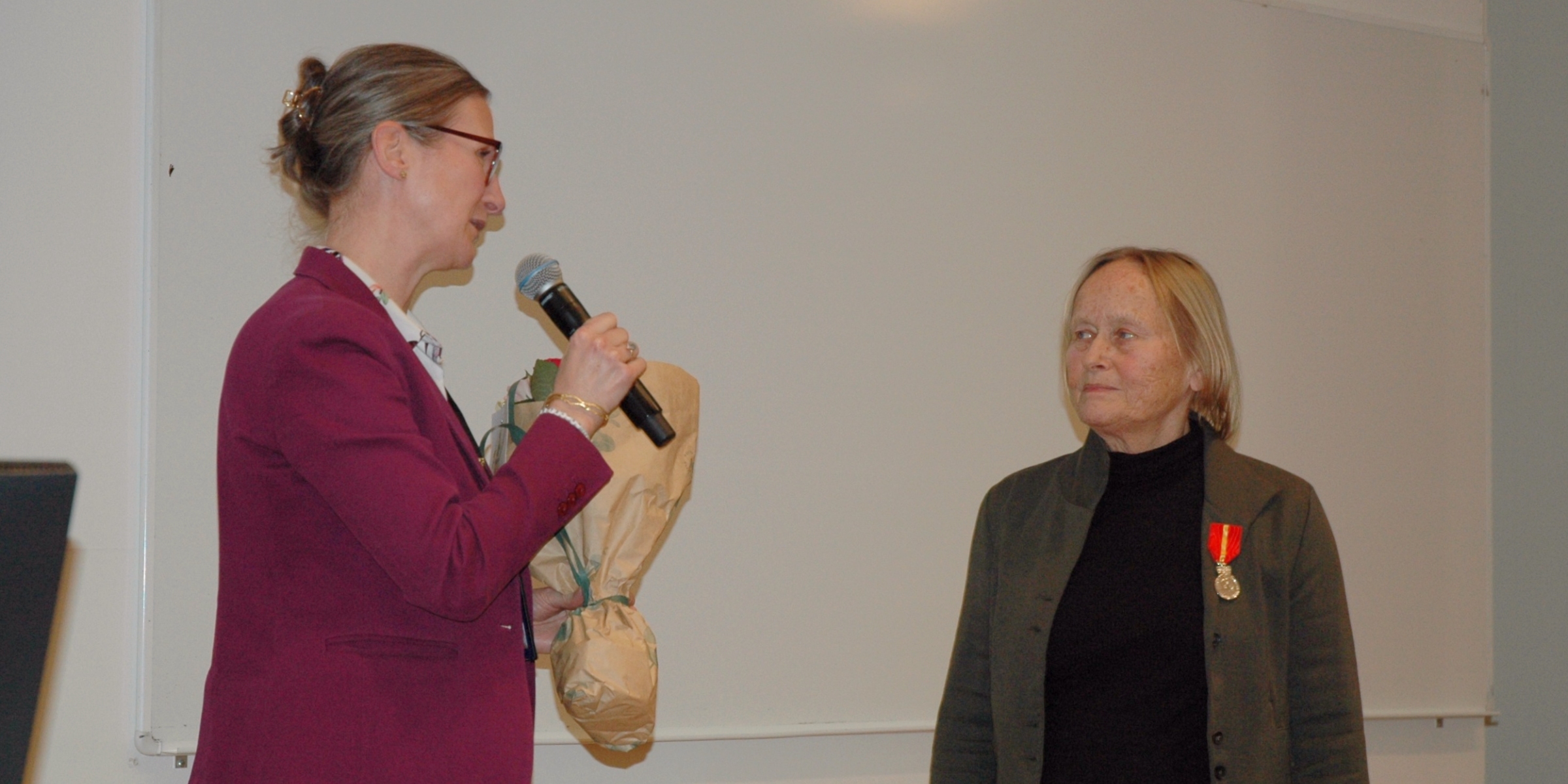 En kvinne med blomsterbukett og mikrofon taler til en kvinne med medalje på brystet