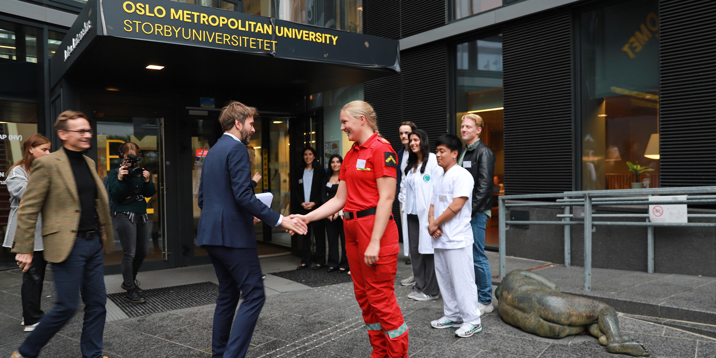 Foto av helseminister Jan Christian Vestre som hilser på en paramedic-student, med flere studenter i rekke som venter utenfor inngangen til Pilestredet 32.