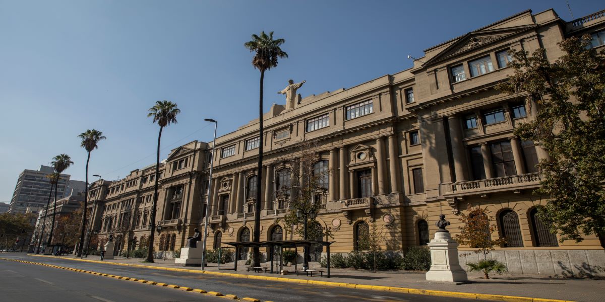 Casa Central campus facade. There are Palm trees at the front of a light brown building of three floors with a Jesus Christ statue at the center of the roof.